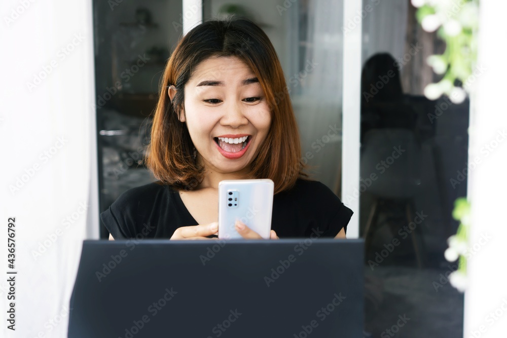 excited Asian woman hand holding mobile phone with smiling face get good news sitting with laptop on desk excited Asian woman hand holding mobile phone with smiling face get good news sitting with laptop on desk