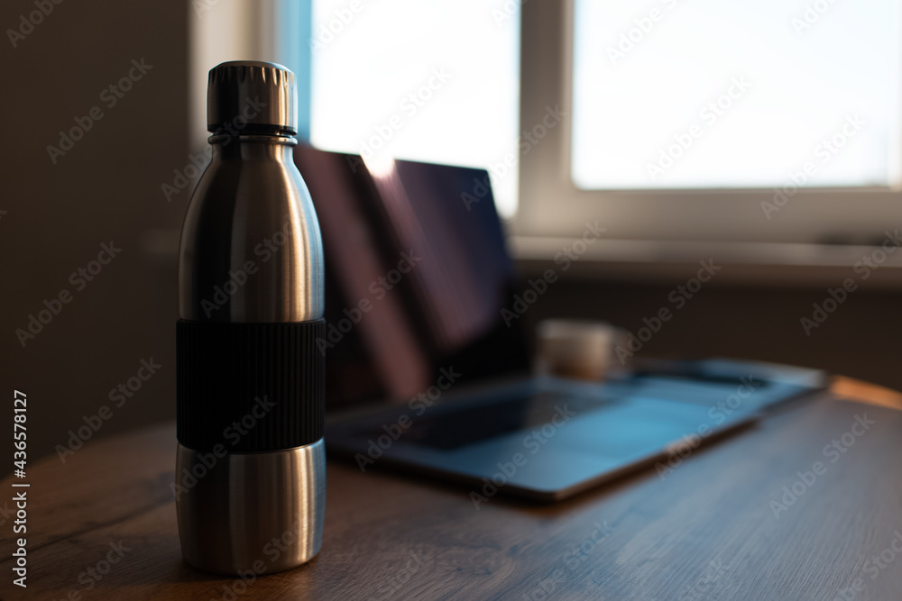 Close-up of steel reusable thermo water bottle on blurred background of laptop. Workspace concept.