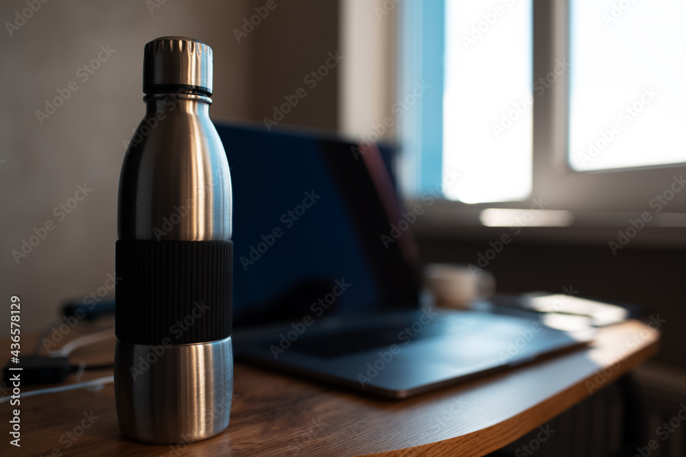 Close-up of steel reusable thermo water bottle on blurred background of laptop. Workspace concept. Dark photo. Close-up of steel reusable thermo water bottle on blurred background of laptop. Workspace concept. Dark photo.