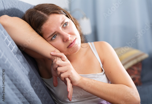 Young sad woman sitting on sofa at home deep in thoughts, feels tired