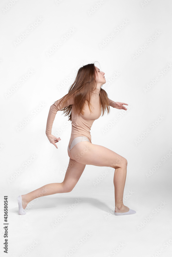 Fototapeta premium Young slender girl, with long hair, in a beige bodysuit, dancing in the white studio. motion blur