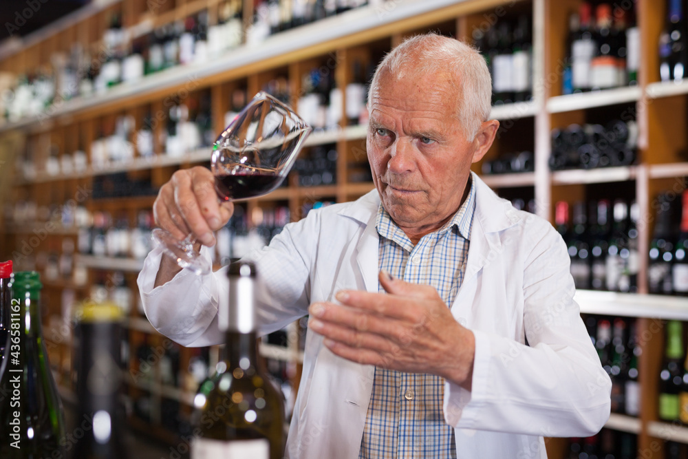 Confident elderly male winemaker holding glass of red wine, checking it in wine store..