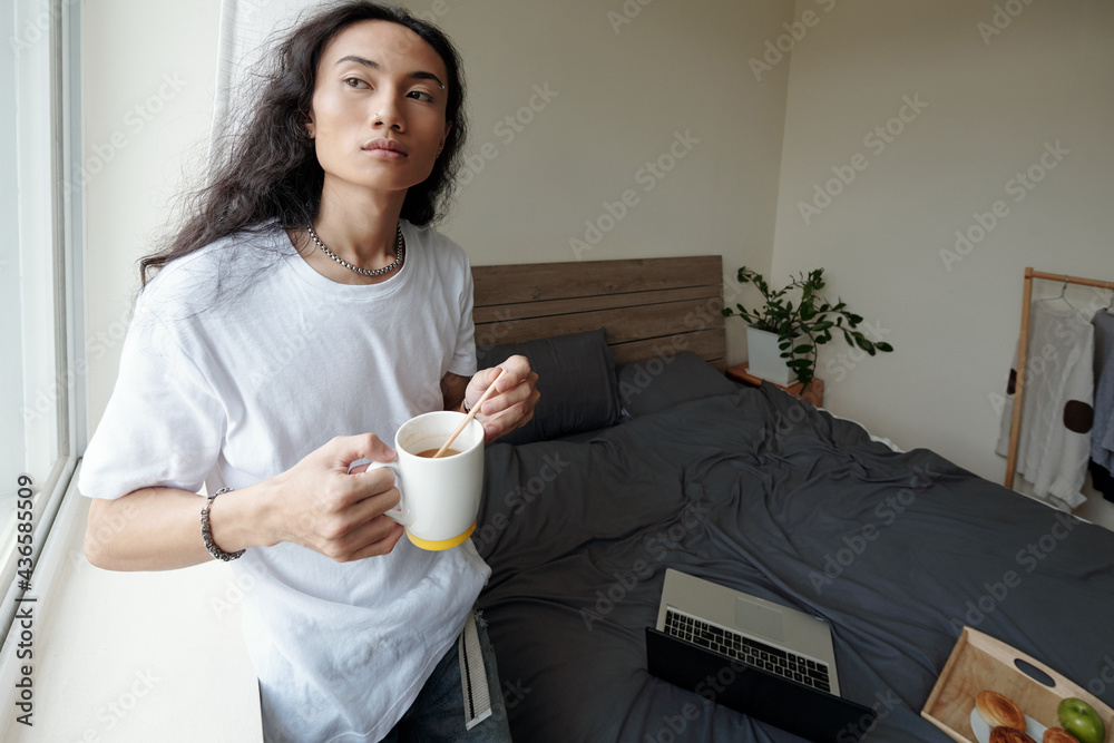 Pensive young Vietnamese man in wavy hair standing at window and stirring sugar in coffee cup, he working remotely at home Pensive young Vietnamese man in wavy hair standing at window and stirring sugar in coffee cup, he working remotely at home