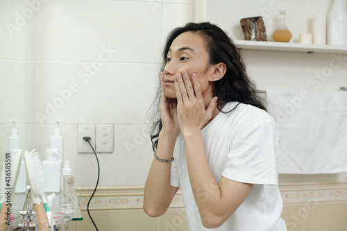 Handsome young Asian man with black wavy hair massaging cheeks while washing face in front of mirror in bathroom