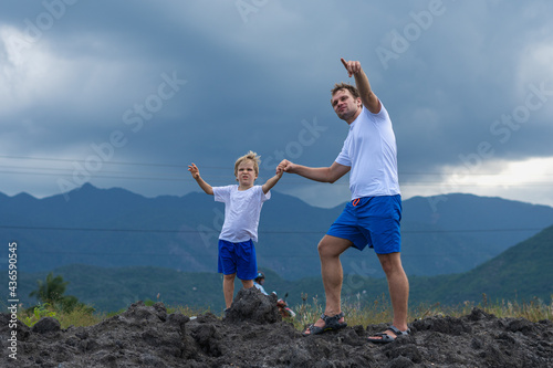 Father son or adult younger brother walk. Cloudy sky before rain, mountain background. Man show points finger into distance, serious boy look far away. Solve problems together. Natural education