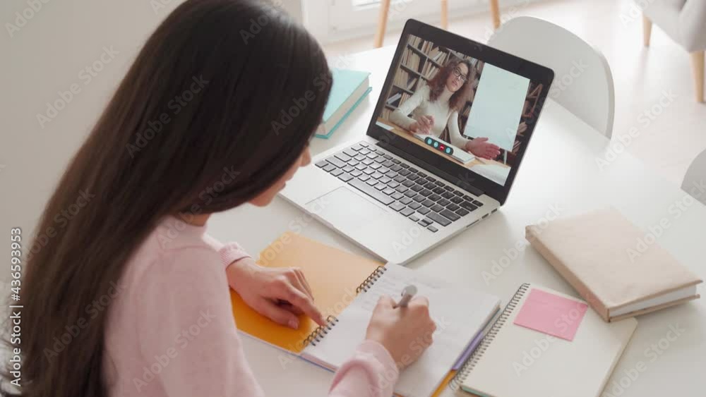 Over shoulder view of cute indian hispanic arab teenage girl having video lesson e learning at home using pc laptop computer writing notes. Remote distant school education concept.