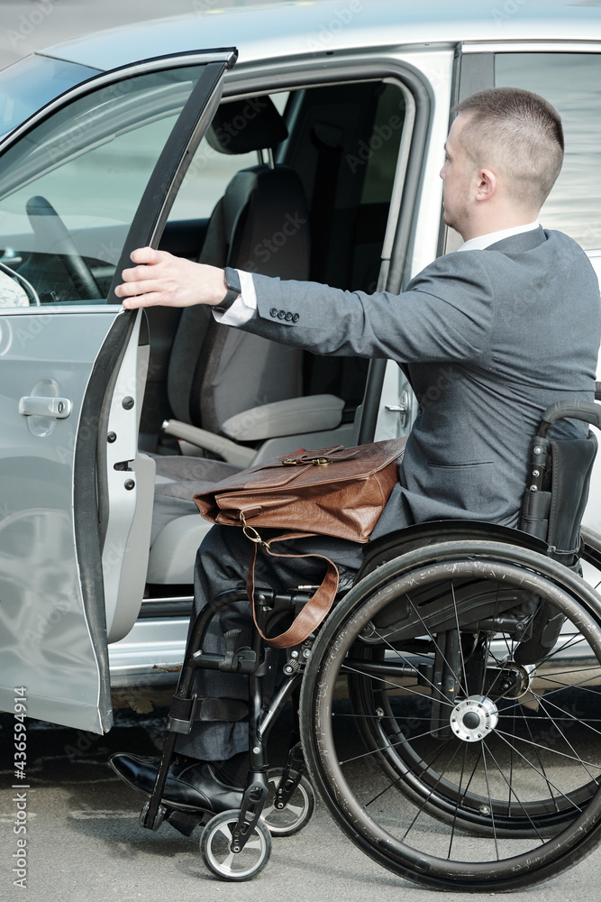 Businessman in formalwear sitting in wheelchair and opening door of his car to get inside