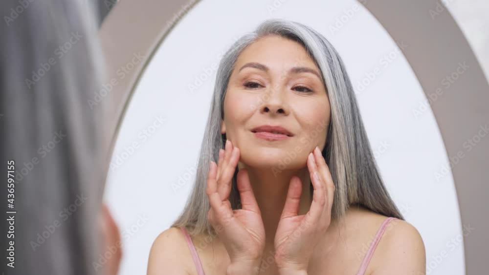 Senior older middle aged Asian woman with grey hair with radiant face with perfect skin looking at mirror smiling to her reflexion touching massaging skin. Ads of senior dry skin care treatment. Senior older middle aged Asian woman with grey hair with radiant face with perfect skin looking at mirror smiling to her reflexion touching massaging skin. Ads of senior dry skin care treatment.