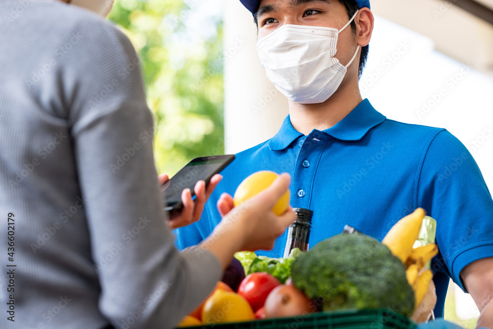 Fototapeta premium Hygienic Asian deliveryman wearing face mask while delivering groceries to customer at home, food delivery in the time of pandemic concept