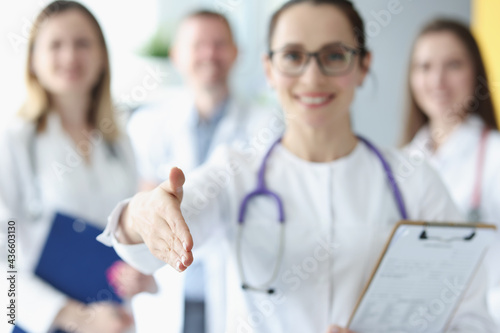 Woman doctor stretches out her hand for greetings against background of medical colleagues