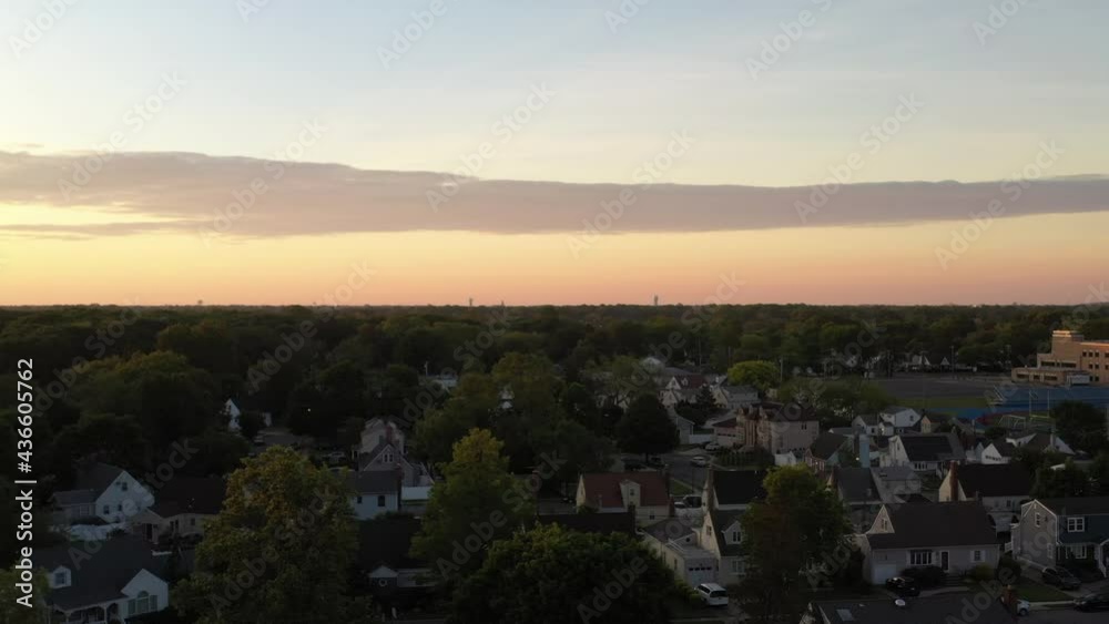 An aerial view over a quiet suburban neighborhood on Long Island, NY at sunrise. The drone camera dolly in and boom up while looking at the colorful sky. The neighborhood below is still. An aerial view over a quiet suburban neighborhood on Long Island, NY at sunrise. The drone camera dolly in and boom up while looking at the colorful sky. The neighborhood below is still.