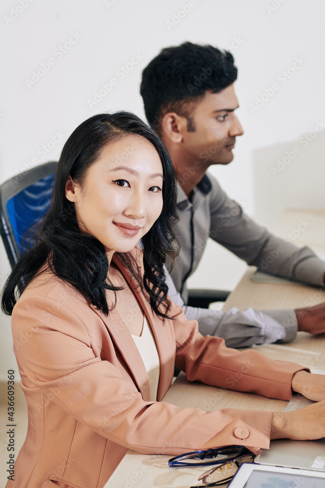 Pretty smiling young woman sitting at office desk next to her colleague and working on laptop Pretty smiling young woman sitting at office desk next to her colleague and working on laptop
