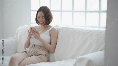 happy asian woman face relaxing using mobile smart phone surfing internet and chat on sofa couch at home. female laying down in living room embarrassed smile when using mobile phone. woman laugh happy