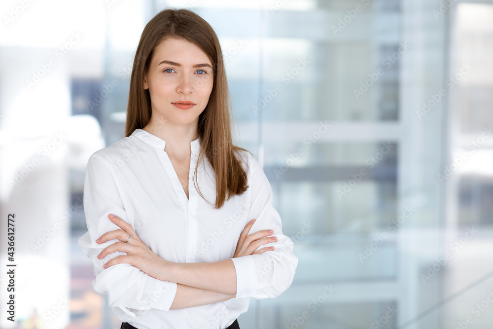 Fototapeta premium Beautiful female specialist with arms crossed standing in modern office and smiling charmingly. Working on design, data analysis, plan strategy. Business people concept