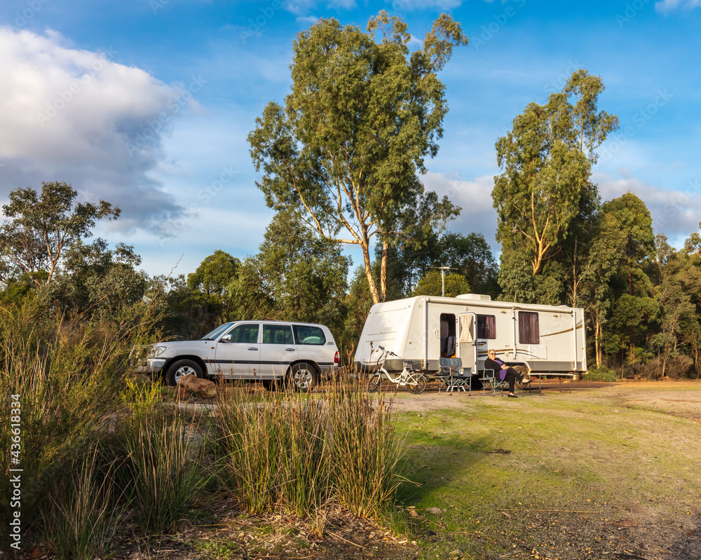 A mature woman sitting outside a large modern caravan and four wheel drive against a background of gum trees under a bright blue sky - obrazy, fototapety, plakaty 