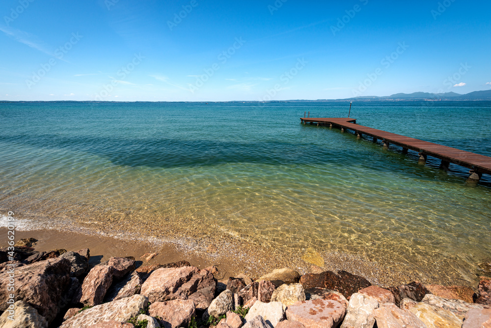 Obraz premium Beautiful beach with a small empty wooden pier on Lake Garda (Lago di Garda) between the small villages of Lazise, Bardolino and Cisano. Tourist resort in Verona province, Veneto, Italy, Europe.