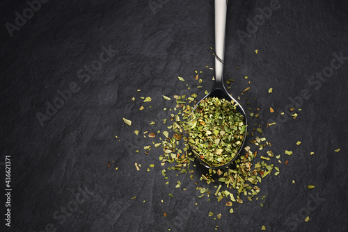 Teaspoon with oregano spice on black slate background.