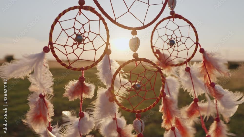 Dream catcher with orange feathers close up rotate in mountains, swaying with light wind slow motion agains blue sky at sunset in summer. Amulet boho style, Indian style. Bright disk of sun, rays sun