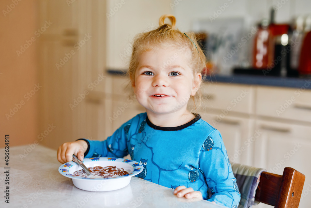 Gorgeous little toddler girl eating healthy cereal with milk for breakfast. Cute happy baby child in colorful clothes sitting in kitchen and having fun with preparing oats, cereals. Indoors at home Gorgeous little toddler girl eating healthy cereal with milk for breakfast. Cute happy baby child in colorful clothes sitting in kitchen and having fun with preparing oats, cereals. Indoors at home