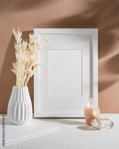 Empty frame with mat, white vase with dried flowers, crystals illuminated by sunlight. Mock up poster with place for your design in the terracotta interior.