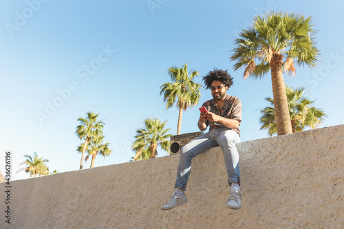 Happy Afro man having fun using mobile smartphone while listening to music with vintage boombox stereo in tropical place during summer vacations