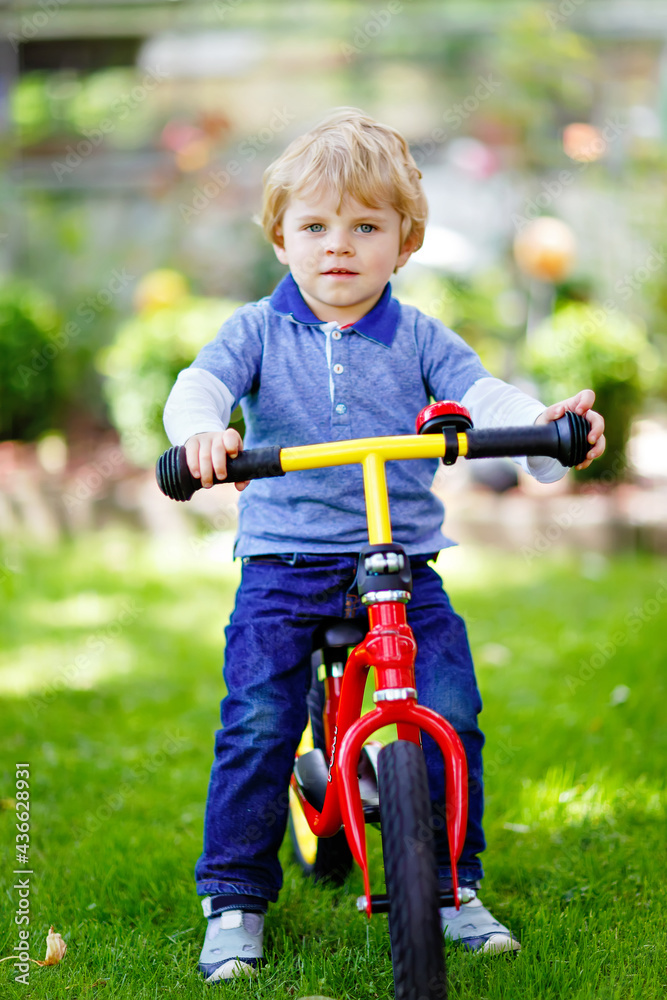 Active blond kid boy in colorful clothes driving balance and learner's bike or bicycle in domestic garden. Toddler child dreaming and having fun on warm summer day. outdoors movement game for children Active blond kid boy in colorful clothes driving balance and learner's bike or bicycle in domestic garden. Toddler child dreaming and having fun on warm summer day. outdoors movement game for children