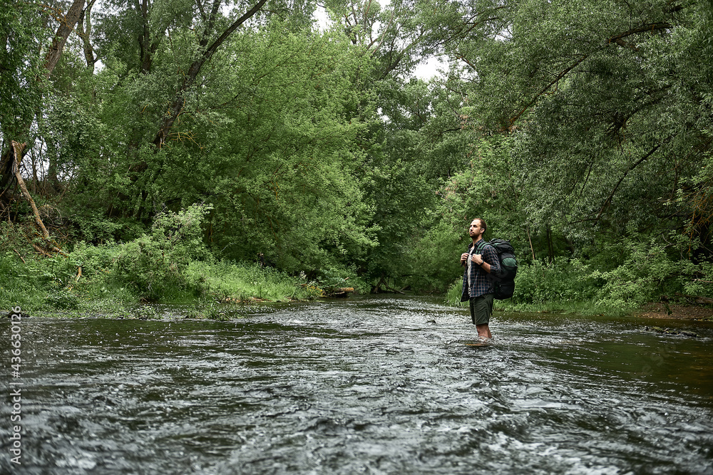 A man in a plaid shirt and green shorts with a large hiking backpack crosses the rive A man in a plaid shirt and green shorts with a large hiking backpack crosses the rive