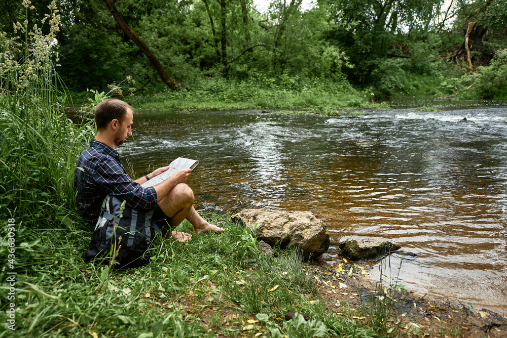 Hiker with a backpack and map sitting on the bank of a river and exploring of the area Hiker with a backpack and map sitting on the bank of a river and exploring of the area