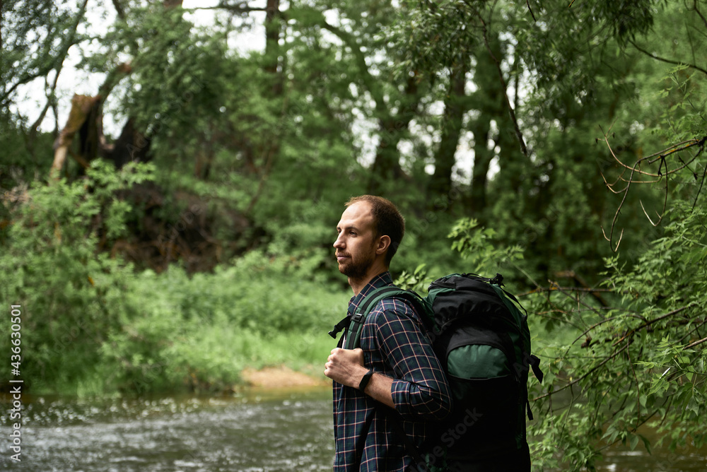 The man with a large hiking backpack standing on the bank of the river and enjoying nature The man with a large hiking backpack standing on the bank of the river and enjoying nature