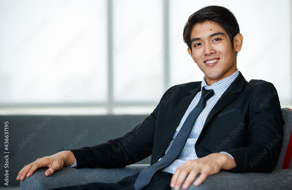 Portrait 20s handsome young successful Asian businessman wearing formal suit with necktie, smiling confidently and intelligently, sitting on comfortable sofa indoor office, posing, looking at camera. Portrait 20s handsome young successful Asian businessman wearing formal suit with necktie, smiling confidently and intelligently, sitting on comfortable sofa indoor office, posing, looking at camera.