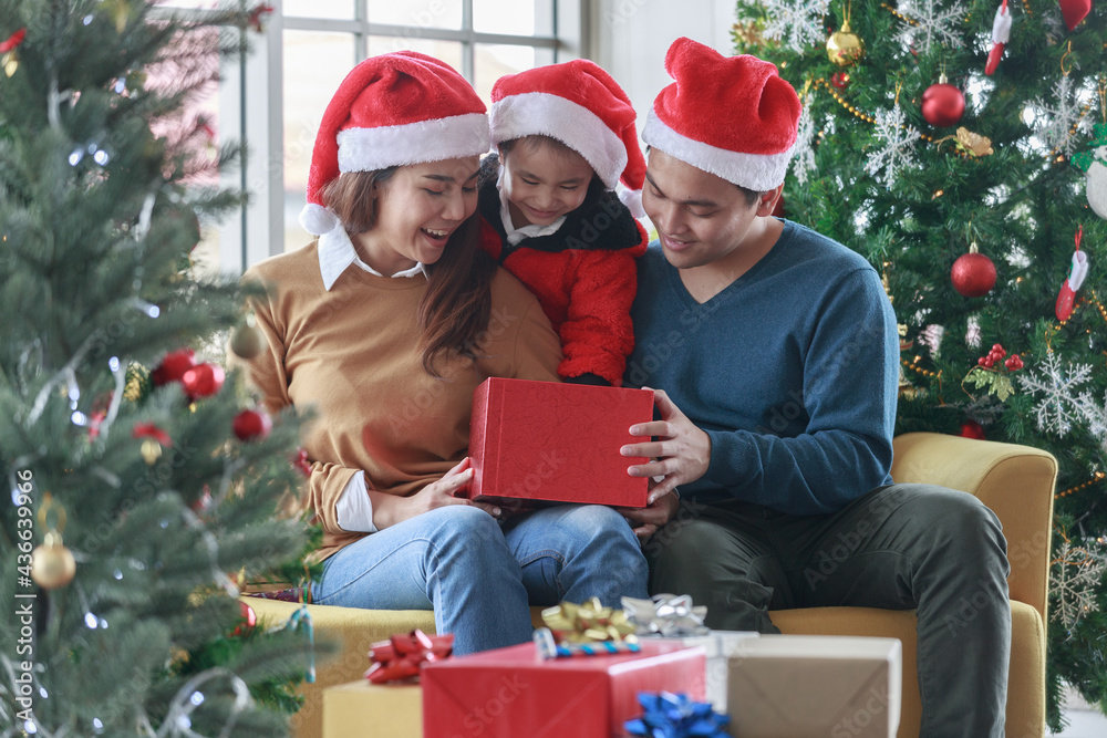Asian happy family and little girl open magic gift box with Christmas tree in living room that decorated for Christmas festival. People Santa hats sitting On the couch. Concept Christmas gift box. Asian happy family and little girl open magic gift box with Christmas tree in living room that decorated for Christmas festival. People Santa hats sitting On the couch. Concept Christmas gift box.