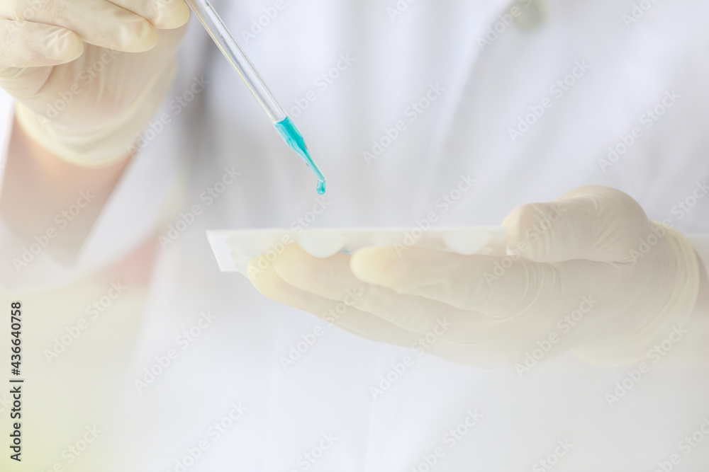Closeup shot of scientist wears white lab coat and rubber gloves hands holding orange red reagent in glass dropper dropping into plastic plates for experiment in laboratory with blurred background Closeup shot of scientist wears white lab coat and rubber gloves hands holding orange red reagent in glass dropper dropping into plastic plates for experiment in laboratory with blurred background