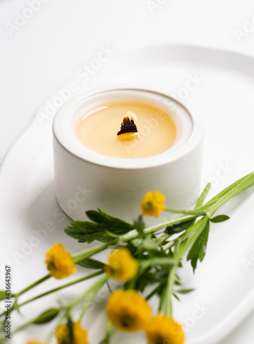 White burning candle with wooden wick and yellow flowers on white tray