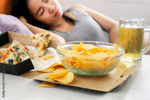 Asian woman sleep after eating junk food with pizza, potato chips and glass of beer on desk, bad habit, unhealthy lifestyle concept photo