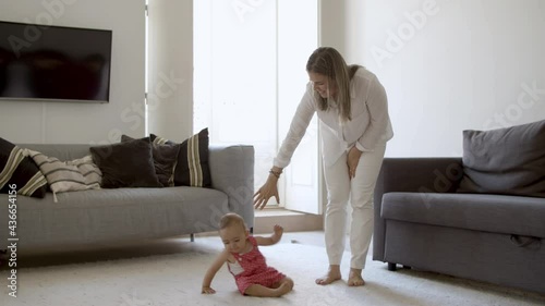 Happy baby in red dungarees crawling on floor. Caucasian mom in casual clothes standing next to daughter, laughing, extending hand to girl to help. Family time, motherhood concept.