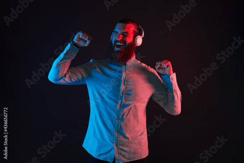 Photo of young bearded hipster man in casual listening music with hands up over dark background