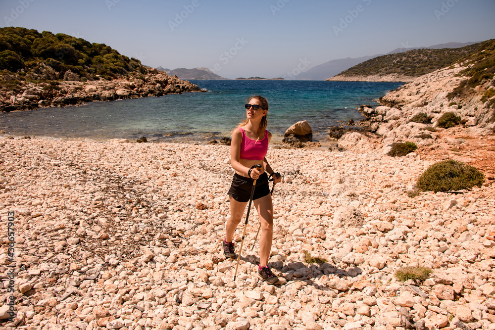 young attractive woman in sunglasses with trekking poles walks along the rocky beach young attractive woman in sunglasses with trekking poles walks along the rocky beach