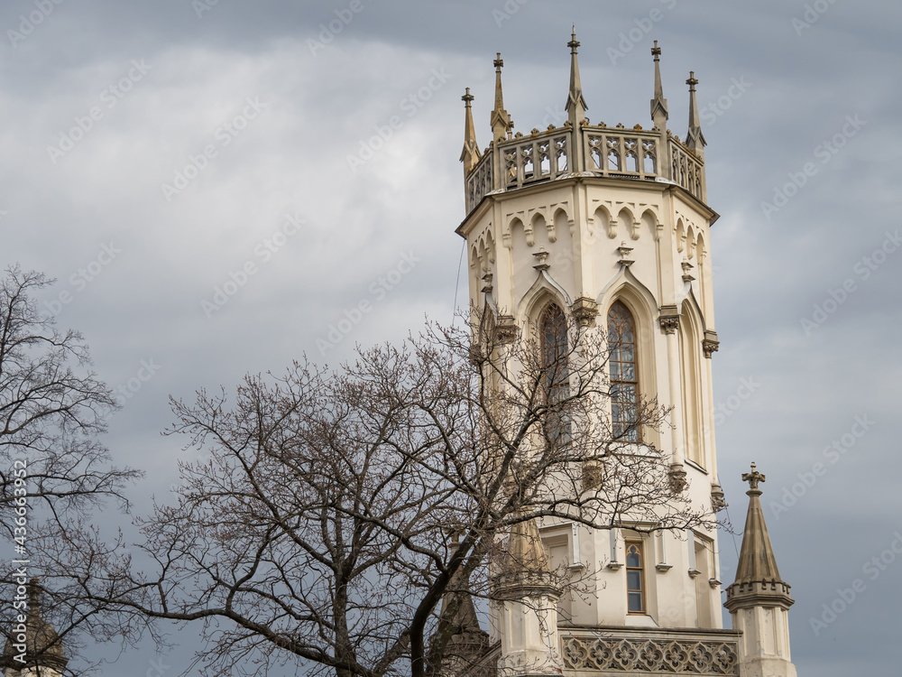 Close-up of the train station tower and bare trees against the sky with clouds. View of the old tower of the Gothic style at the station of New Peterhof. Pseudo-gothic. Beautiful architecture