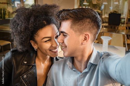 Smiling romantic interracial couple taking selfie sitting in restaurant.