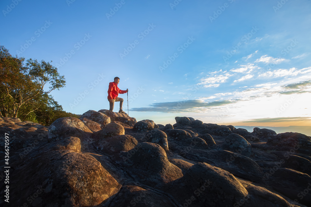 Obraz premium One man in a red jacket with a trekking stick is walking on a rocky hilltop. Sunset time There is a beautiful sky.
