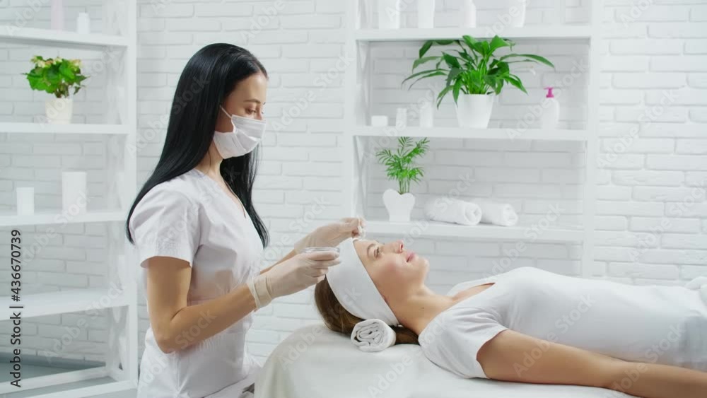 Side view of young brunette beautician in protective mask applying facial mask for young beautiful woman in beauty salon. Concept of beauty procedures for improvements face skin. Side view of young brunette beautician in protective mask applying facial mask for young beautiful woman in beauty salon. Concept of beauty procedures for improvements face skin.