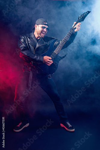Full size photo of brutal bearded Heavy metal musician in leather jacket and sunglasses is playing electrical guitar very emotionally. Shot in a studio on dark background with smoke