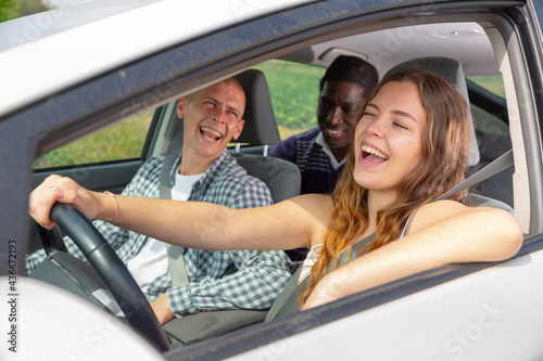 Cheerful young woman driving car with laughing male friends in passenger seats. Friendly road trip and travel concept.. © JackF
