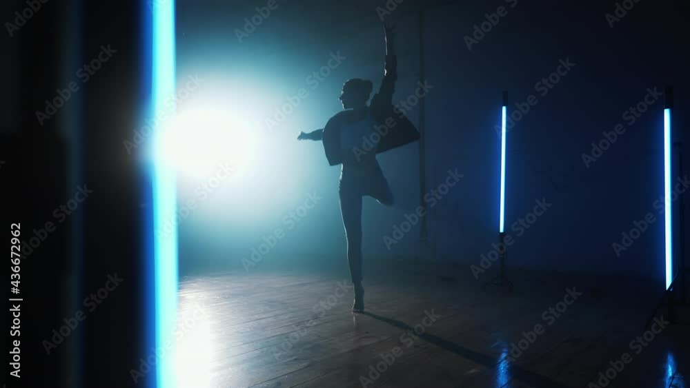Young woman in jeans and jacket practicing in contemporary dance. Studio with blue lamps. Attractive modern ballerina dancing choreography, spinning around. Slow motion.