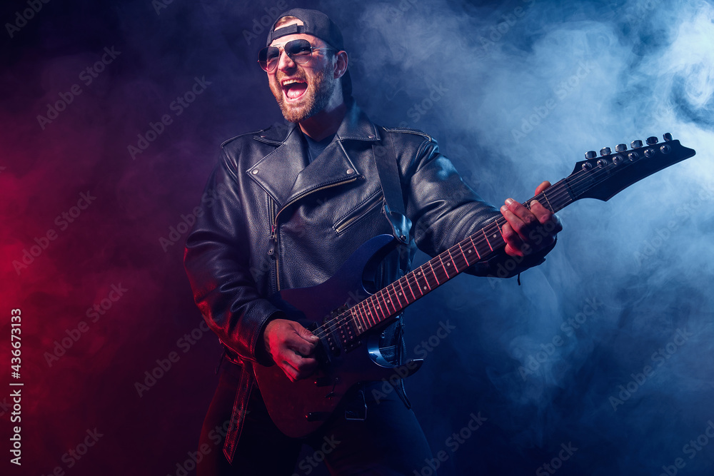 © zamuruev - Brutal bearded Heavy metal musician in leather jacket and sunglasses is playing electrical guitar. Shot in a studio on dark background with smoke