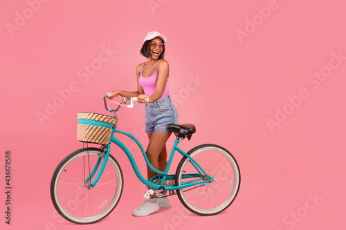 Fototapeta Naklejka Na Ścianę i Meble -  Charming young black lady in summer wear standing with classic bicycle and looking at empty space, pink background