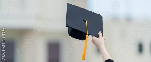 A young beautiful Asian woman university graduate in graduation gown and mortarboard holds a degree certificate stands in front of the university building after participating in college commencement