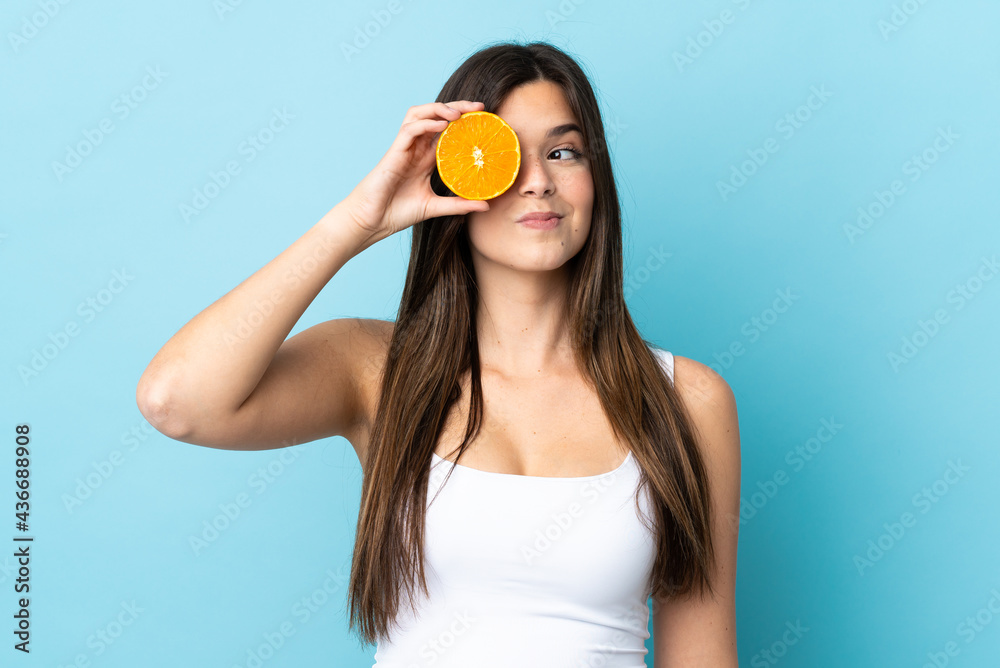 Teenager Brazilian girl over isolated blue background holding an orange Teenager Brazilian girl over isolated blue background holding an orange