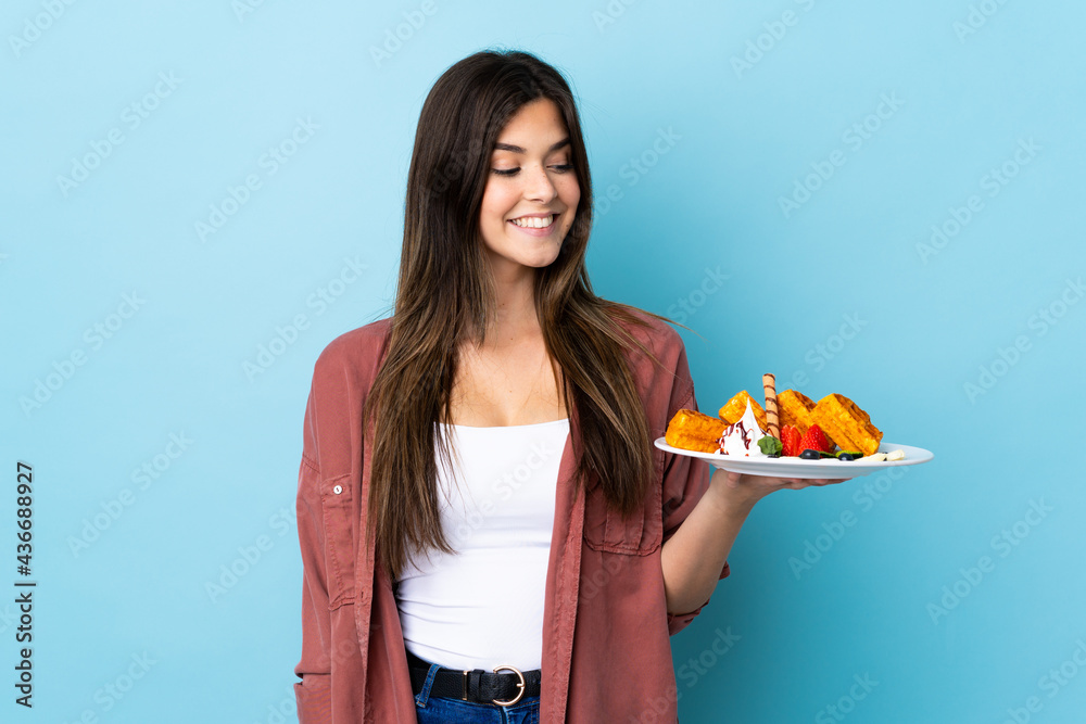 Teenager Brazilian girl holding waffles over isolated blue background with happy expression Teenager Brazilian girl holding waffles over isolated blue background with happy expression