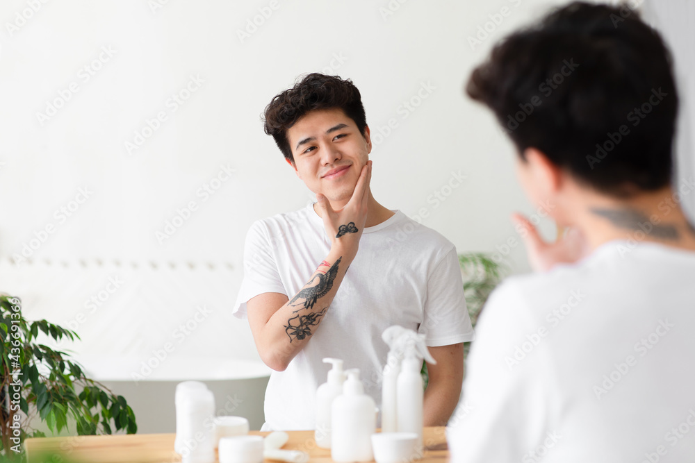 Cheerful asian young guy with tattoos in white t-shirt touches cheek, enjoys clean face Cheerful asian young guy with tattoos in white t-shirt touches cheek, enjoys clean face